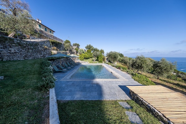 A pool with a stone border, a wooden deck, and a stone wall with a staircase leading to a building.