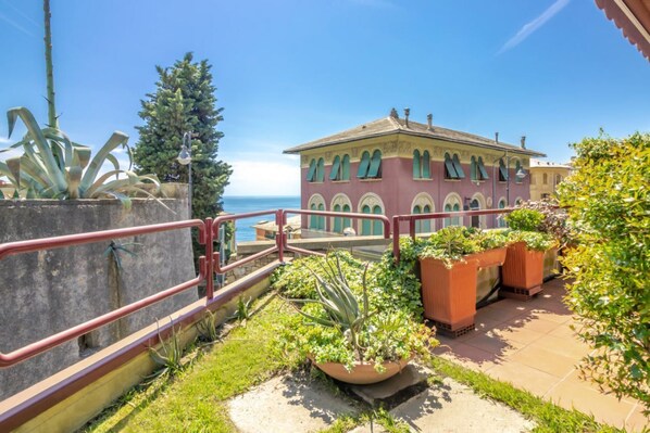 A building with a balcony overlooking a body of water, surrounded by potted plants and a clear sky.