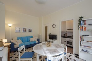 A living room with a sofa, a dining table with chairs, a TV, a bookshelf, and a kitchen area.