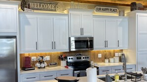 Una cocina moderna con armarios blancos, un refrigerador de acero inoxidable, un microondas y una estufa. Hay taburetes de bar y un letrero que dice “BIENVENIDOS”.