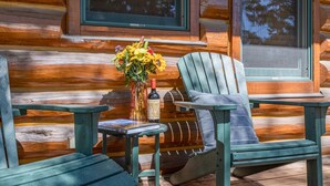 Eine Holzhütte mit einer Terrasse, zwei Adirondack-Stühlen, einem kleinen Tisch und einer Vase mit Blumen.