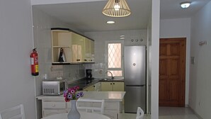 A compact kitchen with white cabinetry, a stainless steel refrigerator, a microwave, and a sink. There's a small dining table with chairs and a vase of flowers. A fire extinguisher is mounted on the wall.