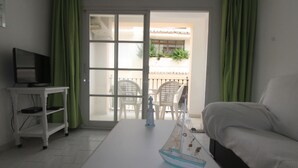 A hotel room with a bed, a small table with a model ship, a TV on a stand, and a sliding glass door leading to an outdoor area with chairs and plants.