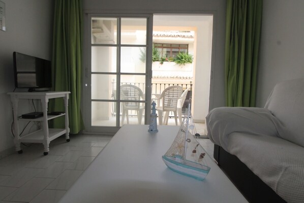 A hotel room with a bed, a small table with a model ship, a TV on a stand, and a sliding glass door leading to an outdoor area with chairs and plants.