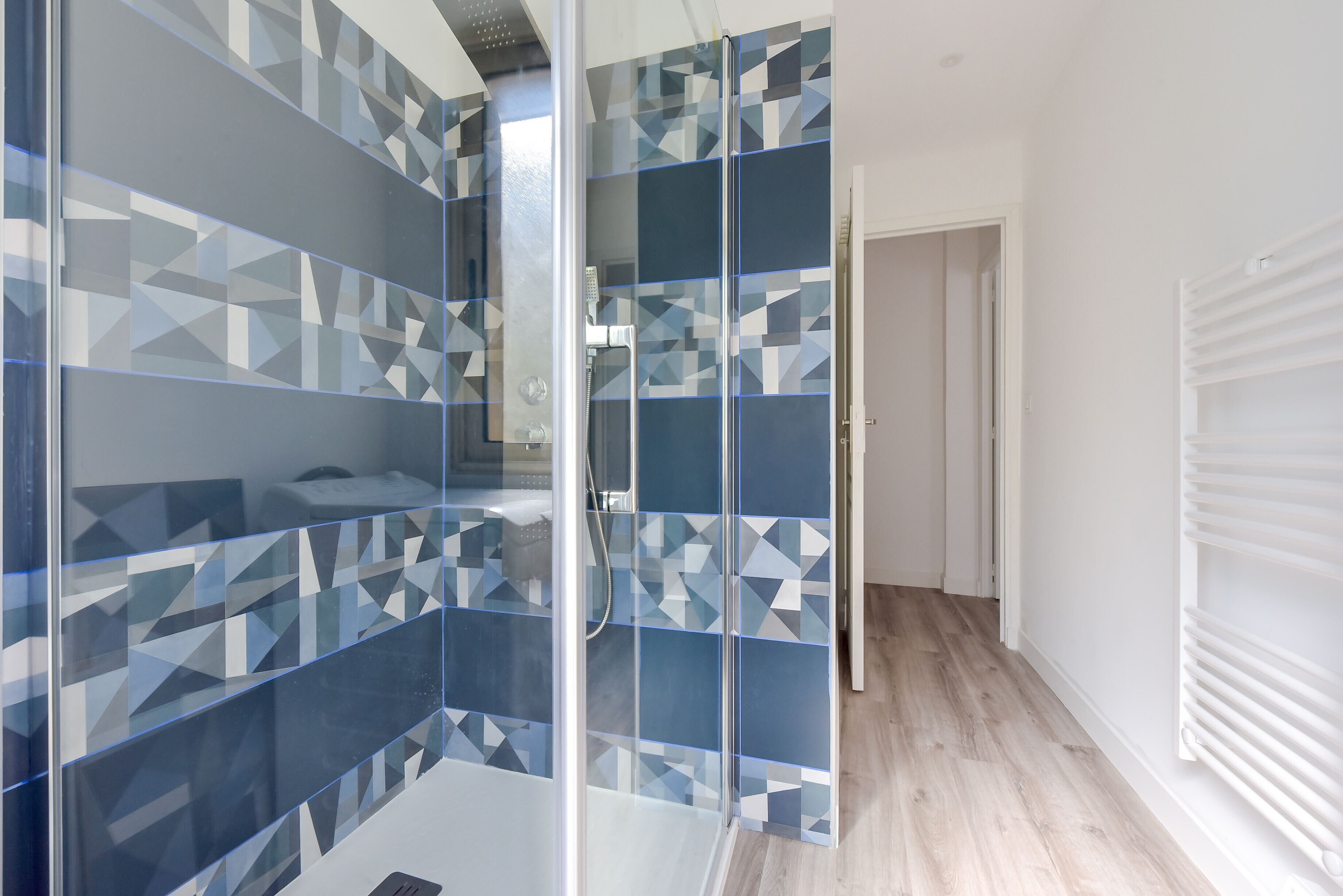A modern bathroom with geometric patterned tiles, a glass shower enclosure, and a white radiator.