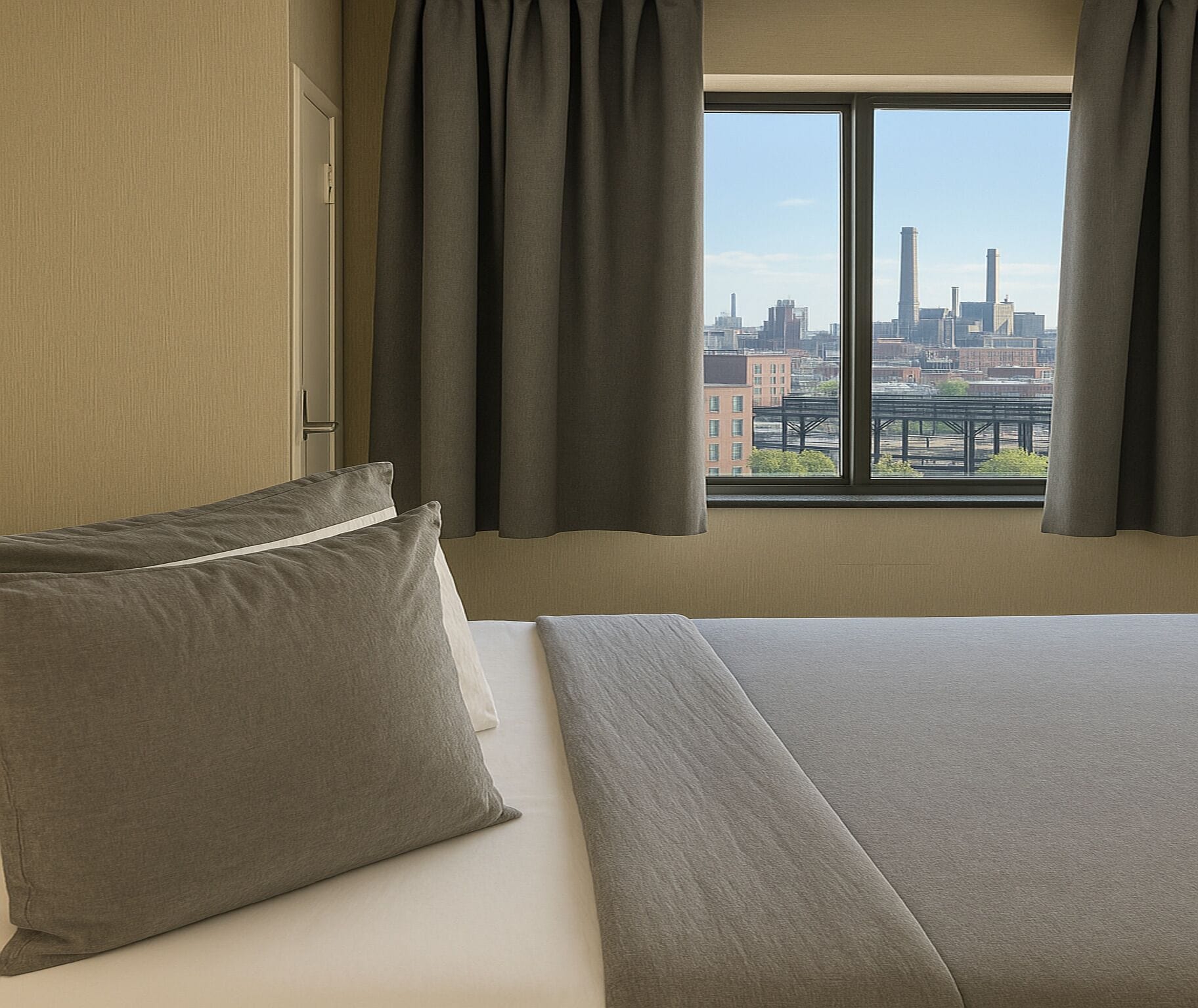 Habitación de hotel con una cama grande, vista a un paisaje urbano por la ventana y una puerta a la izquierda.