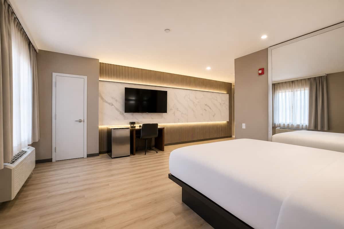 A modern hotel room with a large bed, a desk with a chair, a flat-screen TV mounted on a marble wall, and a window with curtains.