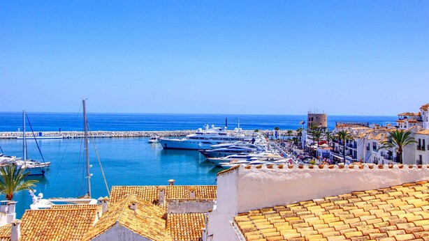 A coastal marina with boats docked, a clear blue sky, and buildings with tiled roofs. - 1079 - FRONTLINE PUERTO BANUS APARTMENT (Marbella)