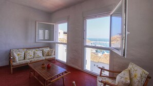 A room with a sofa, a wooden table, and two chairs. There are large windows with a view of buildings and the sea. - 1079 - FRONTLINE PUERTO BANUS APARTMENT (Marbella)