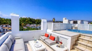 Una terraza en la azotea con piscina, mobiliario blanco y vistas a la ciudad.