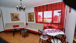 Una habitación acogedora con sofá, una mesita, una mesa de comedor con sillas, una araña y un ventanal con cortinas rojas.
