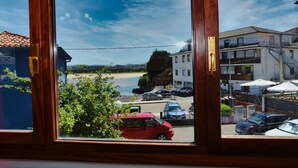 Vista a través de una ventana de un edificio con vista a una playa, coches estacionados y un edificio con terraza.