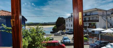 A view through a window of a building overlooking a beach, parked cars, and a building with outdoor seating.