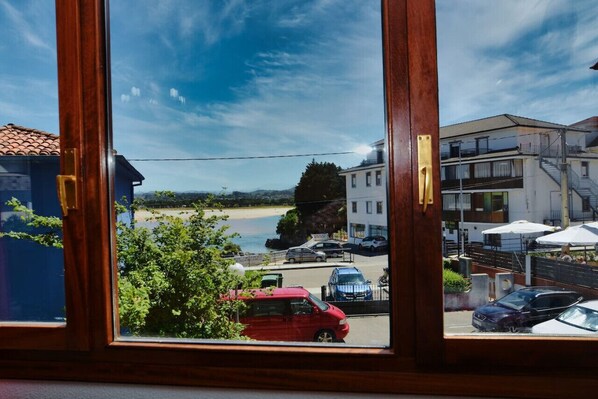 A view through a window of a building overlooking a beach, parked cars, and a building with outdoor seating. - Apto 2 habitaciones con piscina - Isla Playa 5709 (Arnuero)