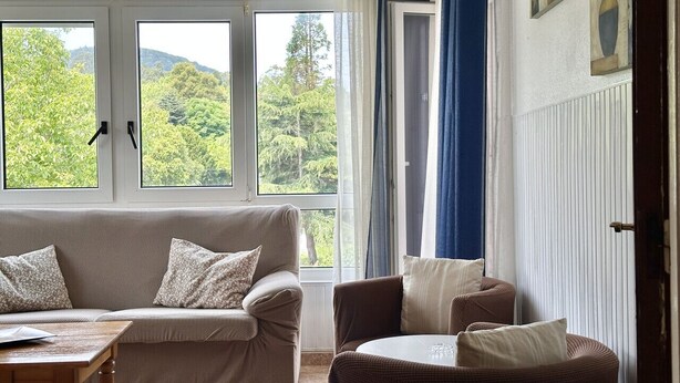 A living room with a beige sofa, a brown armchair, and a wooden coffee table. There are two windows with curtains, and a door with a view of greenery outside. - Apto con jardín compartido - Villa Cancillo 6802 (Noja)