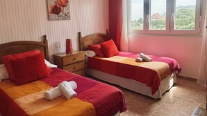 A hotel room with two beds, each with red bedding and white towels, a wooden headboard, a nightstand, and a window with a view of greenery. - Apto con jardín compartido - Villa Cancillo 6802 (Noja)