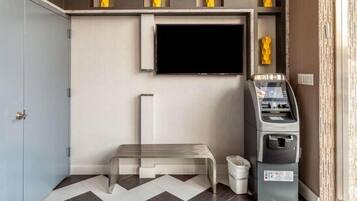Habitación de hotel con televisor de pantalla plana montado en la pared, un banco, un cesto de basura y un cajero automático.
