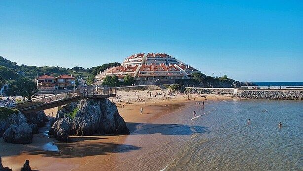 Una spiaggia con un litorale roccioso, un ponte e un edificio con tetti rossi.