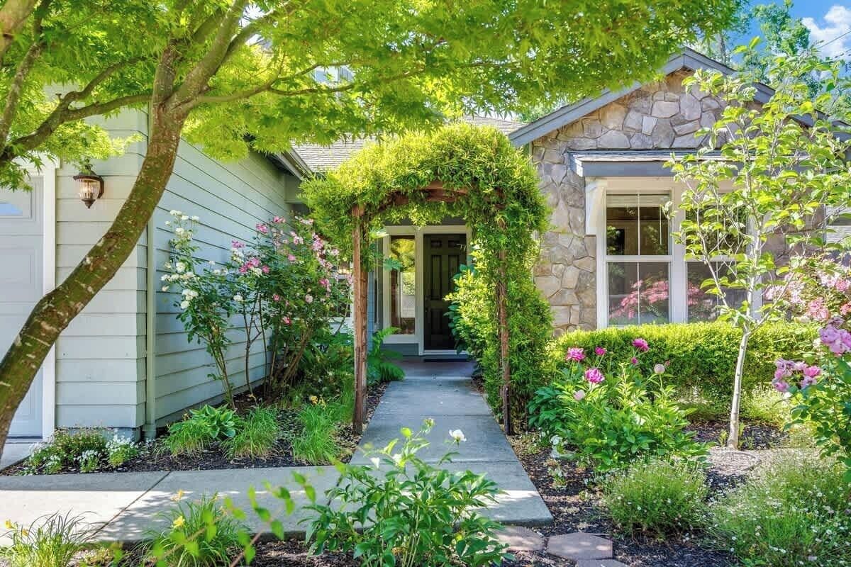 Une maison avec une façade en pierre, un porche abrité et un jardin où poussent différentes plantes et fleurs.