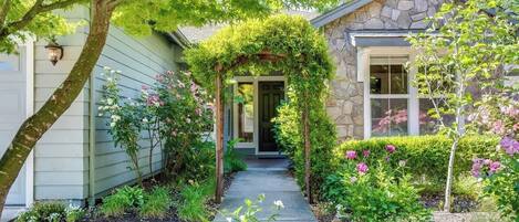 Een huis met een stenen gevel, een overdekte veranda en een tuin met verschillende planten en bloemen.