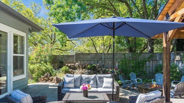 An outdoor patio area with a sofa, wicker chairs, a table with a vase of flowers, and a large umbrella. - Wine Country Harvest Vacation Home (Santa Rosa)