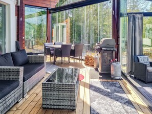 A modern outdoor living area with a glass enclosure, furnished with sofas, a table, and chairs, and a barbecue grill.