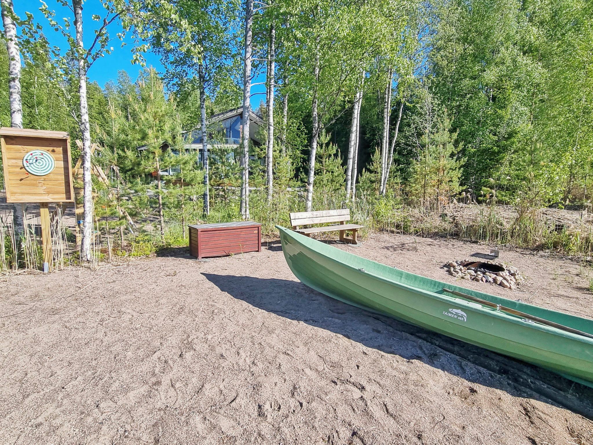 Een kano, een houten bankje en een klein tuinhuisje in een natuurlijke omgeving, met bomen en een gebouw op de achtergrond.