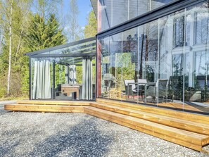 A modern glass house with a wooden deck, outdoor furniture, and a view of the surrounding forest. - Nummentähti by Interhome (Tammela)