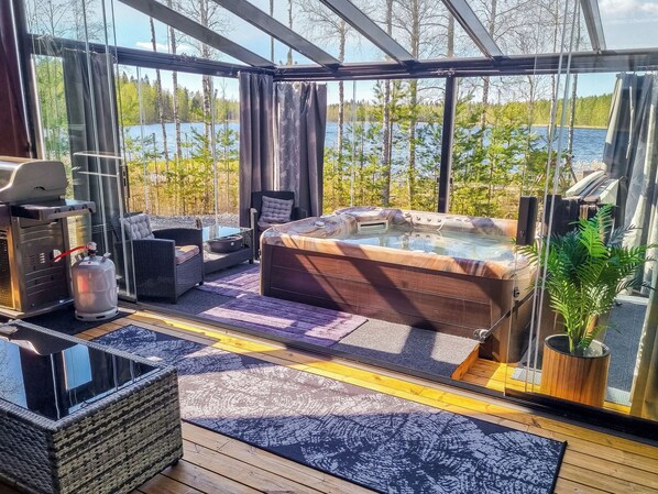 A glass-enclosed room with a hot tub, seating area, and a view of a lake.