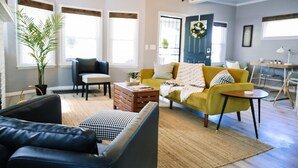 Un salon avec un canapé jaune, un divan en cuir noir et une table basse en bois. On y trouve également une plante en pot et un lustre.