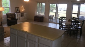 Uma sala de estar ampla com uma grande ilha na cozinha, uma mesa de jantar com cadeiras, um ventilador de teto e vista para o exterior através de janelas grandes.