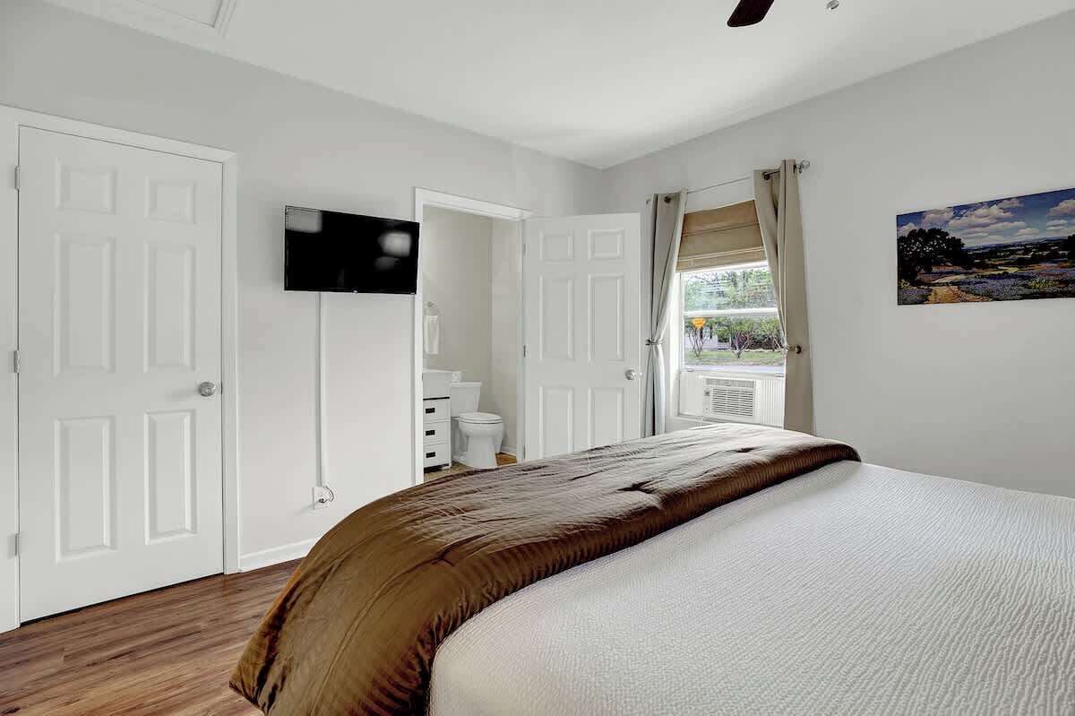 Um quarto com cama, televisão na parede, janela com cortinas e porta.