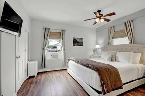 A bedroom with a bed, a ceiling fan, a window with curtains, a wall-mounted TV, and a door. - Bluebonnet House-Deck w/fire table (Fredericksburg)