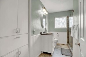 A bathroom with a white vanity, a sink, a mirror, a toilet, and a shower area. - Bluebonnet House-Deck w/fire table (Fredericksburg)