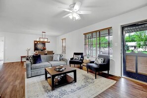 A living room with a grey sofa, a black armchair, a coffee table, and a ceiling fan. There is a large window with blinds and a view of greenery outside.