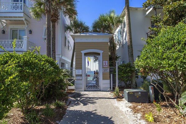 A narrow pathway leading to a building with a gated entrance, surrounded by lush greenery and palm trees.
