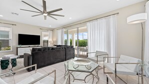 A modern living room with a ceiling fan, a flat-screen TV, a sofa, a glass coffee table, and a dining area with chairs and a lamp.
