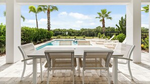 A covered outdoor area with a pool, white furniture, and palm trees.