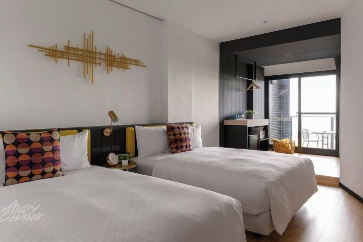 Habitación de hotel con dos camas, una obra de arte en la pared, una mesita con una lámpara y un balcón con una silla.