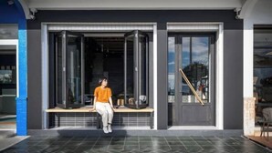 Une personne est assise sur un banc devant un bâtiment doté de grandes portes vitrées et d’un escalier menant à l’intérieur.