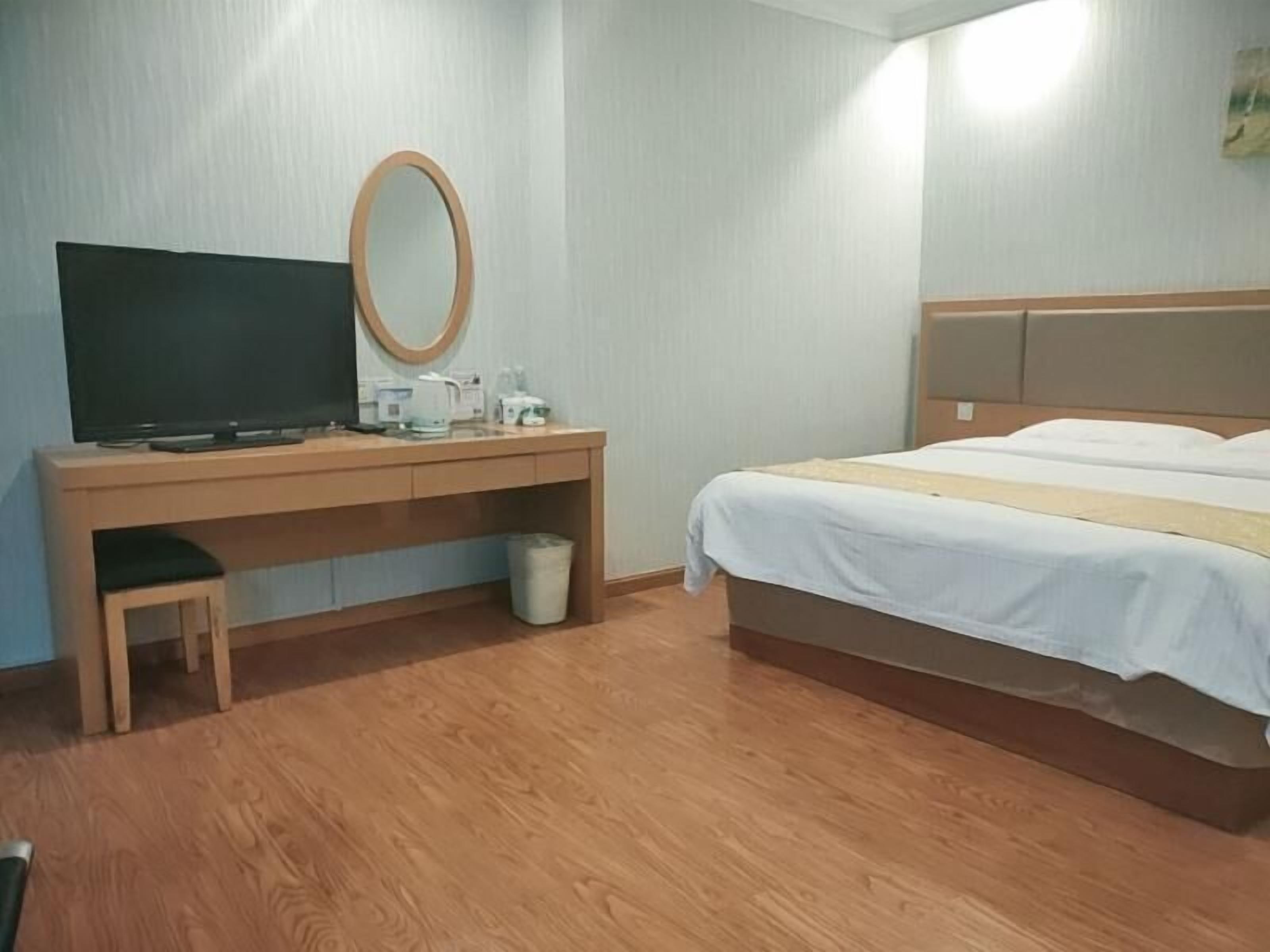 Habitación de hotel con escritorio de madera, televisor, espejo, cama con ropa blanca y un pequeño cubo de basura.