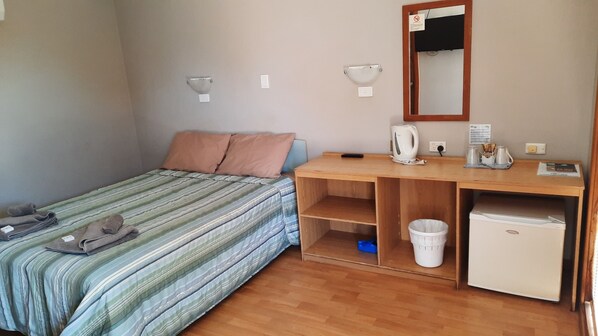 Una habitación pequeña y ordenada con una cama, un escritorio de madera, un espejo, una tetera y un refrigerador pequeño.