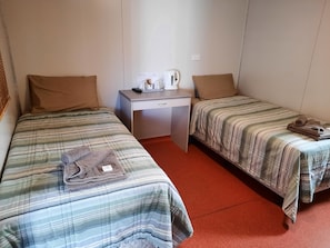 A room with two beds, each with striped bedding, a desk with a kettle and a remote control, and a pair of rolled towels on one bed.