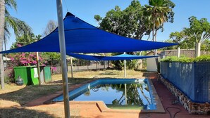 A backyard with a swimming pool, a blue shade sail, and a green trash bin.