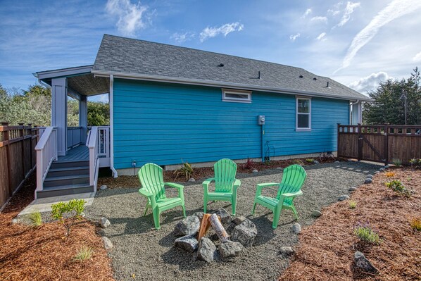 Une maison bleue avec un porche abrité, trois chaises Adirondack vertes, un foyer et une allée gravillonnée.