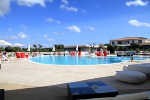 Una piscina con sillas de descanso y sombrillas, rodeada por una valla blanca y palmeras.