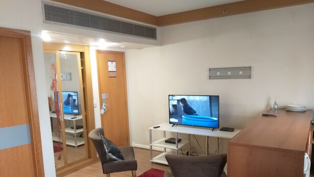 Une chambre d’hôtel avec un téléviseur posé sur un meuble blanc, deux fauteuils gris, un tapis rouge et un bureau en bois.