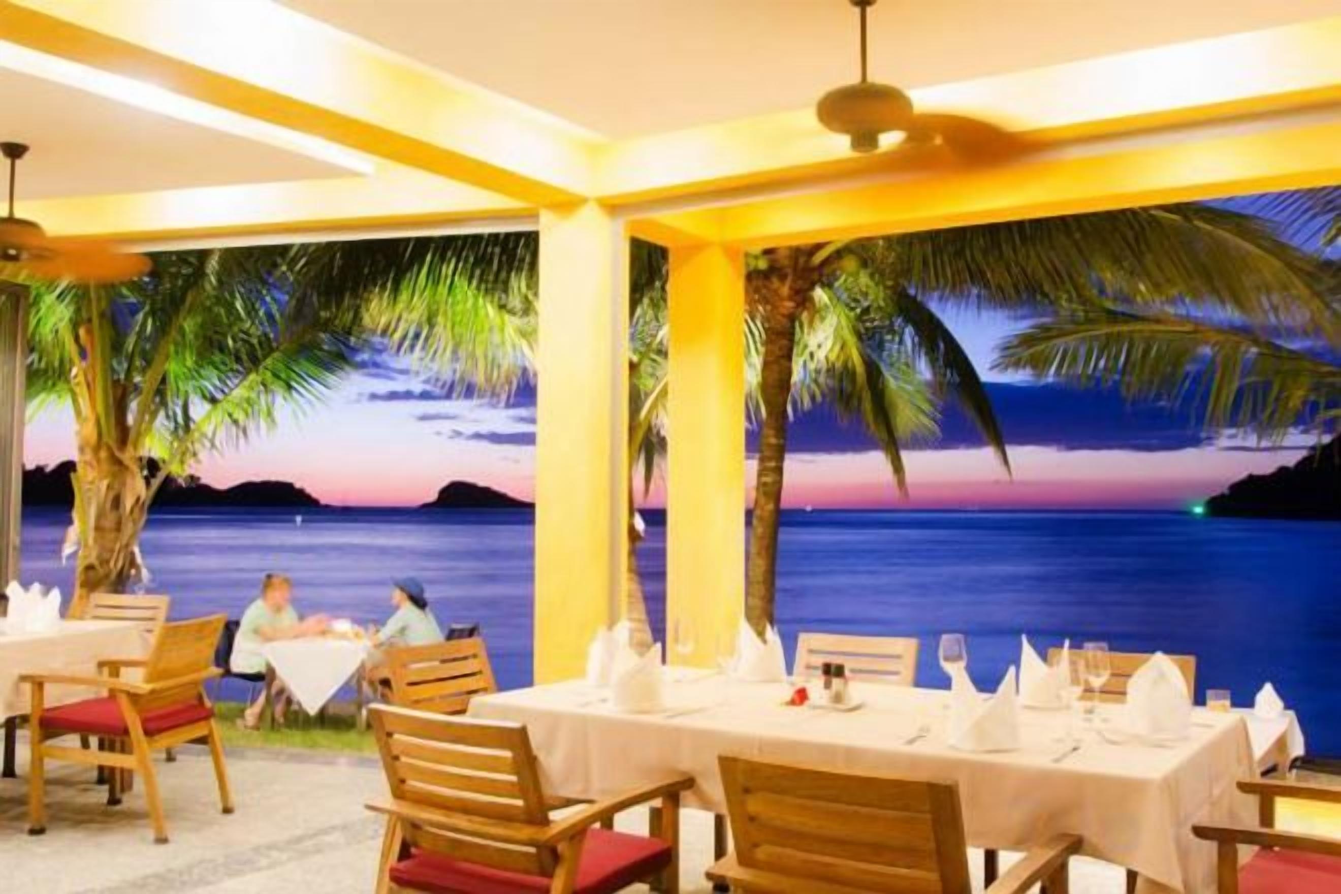A restaurant with tables set for dining, a view of the sea, and palm trees.