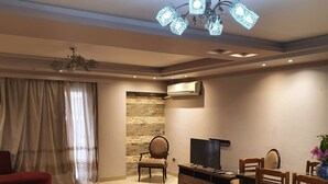 A modern living room with a flat-screen TV, a dining area with wooden chairs, a red sofa, a patterned rug, and a stone accent wall. - Charming 3-Bed Apartment in New Cairo City (New Cairo City)