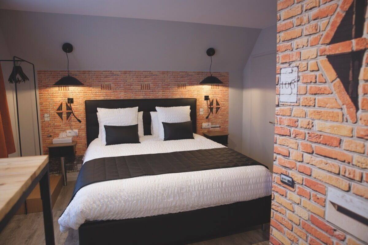 Un dormitorio con pared de ladrillo, cama con ropa blanca, dos lámparas de noche y una mesita de madera.